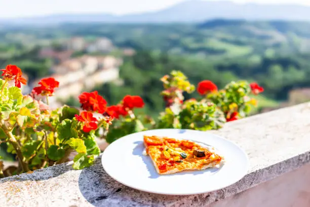 Closeup of pizza slice on white plate on balcony terrace by red geranium flowers outside in Italy with mountain view in Chiusi, Umbria or Tuscany