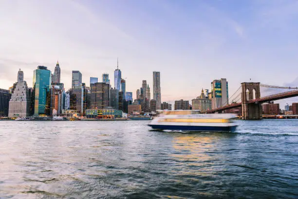 Outdooors view on NYC New York City Brooklyn Bridge Park by east river, cityscape skyline at sunset, dusk, twilight, skyscrapers, buildings, waves, blur, blurry tour boat