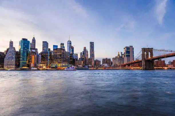 Outdooors view on NYC New York City Brooklyn Bridge Park by east river, cityscape skyline at sunset, dusk, twilight, blue hour, dark night, skyscrapers, buildings, waves, blurred, blur, blurry tour boat