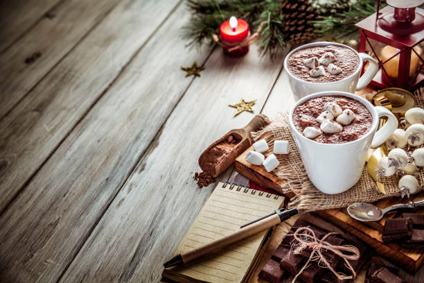 Homemade hot chocolate mugs with marshmallows on rustic wooden table with candlelight and note pad. Christmas themes.
