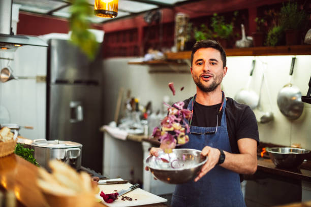 Action portrait of male chef tossing ingredients in bowl Young man preparing fresh food in commercial kitchen, looking at camera, fresh ingredients mid air mixing photos stock pictures, royalty-free photos & images