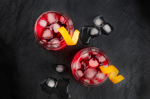 Two vibrant cocktails with campari and orange zest twist garnishes, shot from above with ice cubes on a black background, with copy space