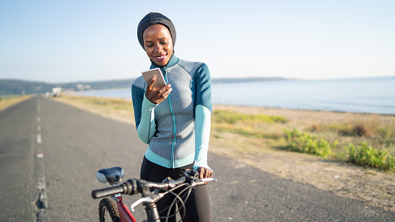 African sportswoman with hijab, with a bike using smart phone on road