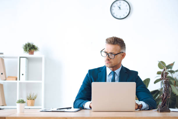 istock handsome lawyer in eyeglasses using laptop and looking away in office 1059944196