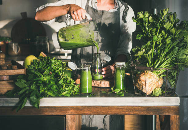 Woman pouring green smoothie from blender to bottle Making green detox take-away smoothie. Woman in linen apron pouring green smoothie drink from blender to bottle surrounded with vegetables and greens. Healthy, weight loss food concept detox stock pictures, royalty-free photos & images