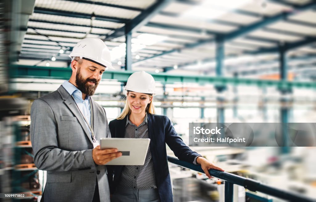 A portrait of an industrial man and woman engineer with tablet in a factory, working. A portrait of a mature industrial man and woman engineer with tablet in a factory, working. Industry Stock Photo