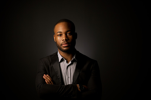 A strong business portrait of a young African businessman with formal wear studio lit against a dark background with arms crossed Cape Town South Africa