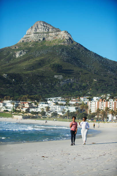 Two young African women walking laughing Having Ice cream on the Camps Bay beach with Lions Head mountain backdrop  Cape Town South Africa