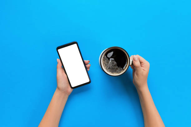 femininas mãos segurando um celular preto com tela branca em branco e a caneca de café. imagem da maquete com espaço de cópia. vista superior sobre fundo azul, plana leigos - text messaging mobile phone teenagers only people - fotografias e filmes do acervo