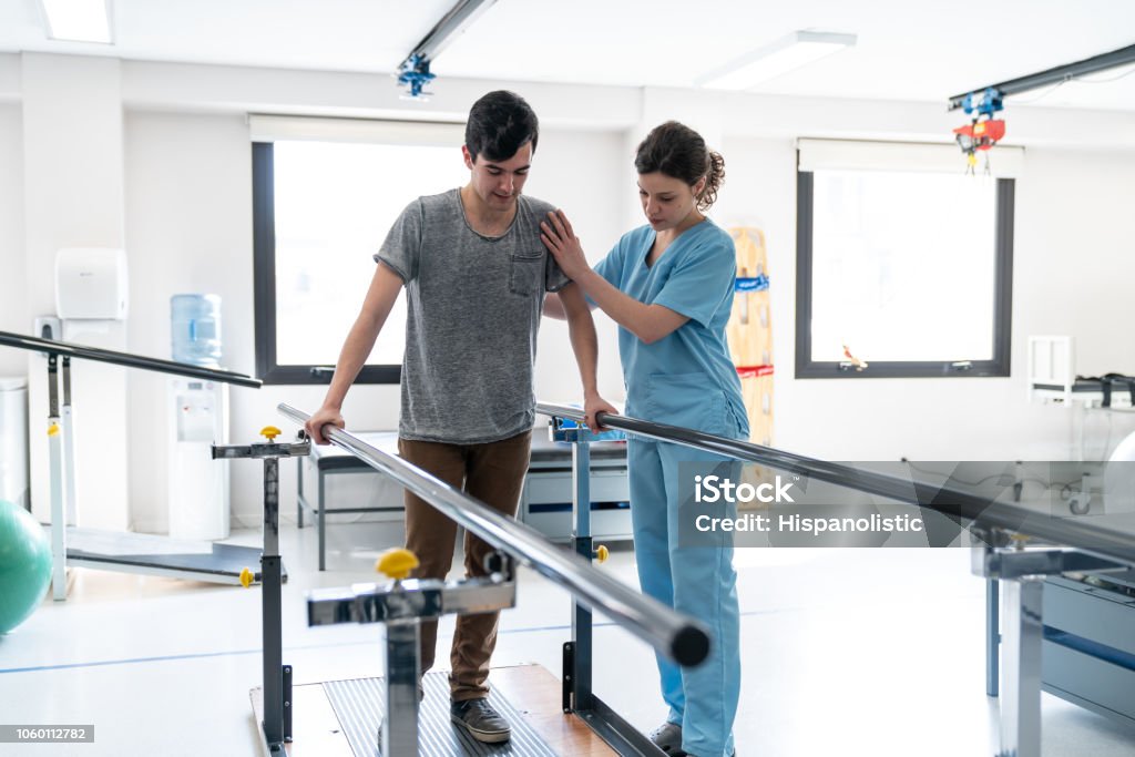 Paziente maschio concentrato in fisioterapia che cammina con l'aiuto di barre parallele e terapista accanto a lui - Foto stock royalty-free di Fisioterapia