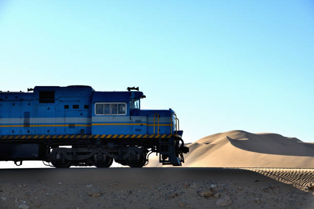 The train is dominant in the photo. Behind the train is a sand dune, against blue sky. The photograph was taken in the Namib Desert, close to Walvis Bay, Namibia, in February 2018.