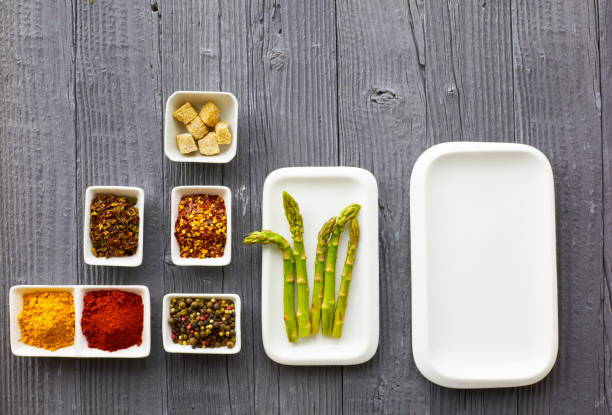 Horizontal image of fresh vegetables placed on a wooden tabletop. Asparagus, bell peppers, mushrooms, tomatoes and one lemon sit in the bright window light. There is a lovely reflection of the vegetables on the table with a bright white wall and kitchen window in the background.