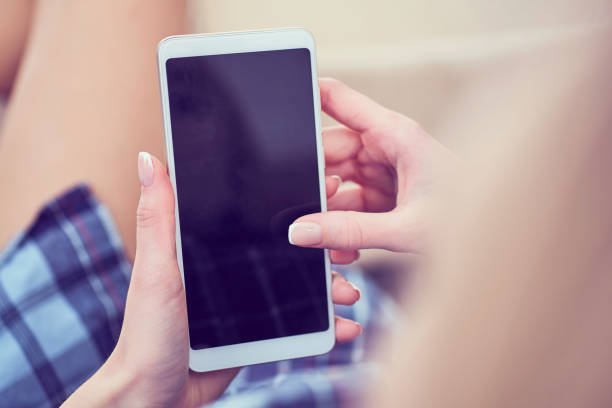 Back view of a woman hands using smart phone with blank screen close up. White phone with a black screen in the hands of a young girl lying on the floor with her feet on the couch