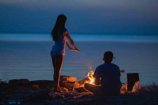 the couple resting on the shore near the bonfire. evening night time - bonfire beach fire barbecue imagens e fotografias de stock