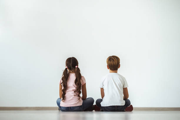 la ragazza e un ragazzo seduti sul pavimento sullo sfondo bianco della parete - rear view family isolated child foto e immagini stock