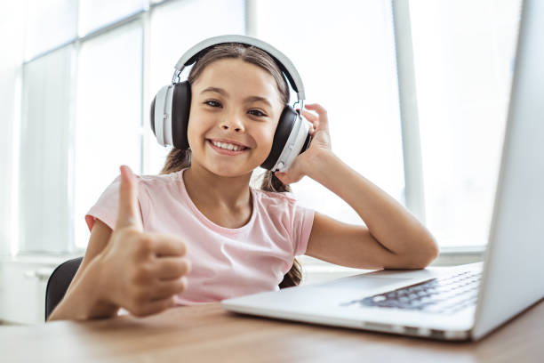 szczęśliwa dziewczyna w słuchawkach z laptopem trzyma kciuki przy stole - happy kid audio zdjęcia i obrazy z banku zdjęć