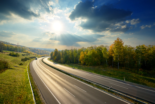 Empty asphalt highway with truck arriving from a distance under the radiant sun and dramatic clouds in the countryside in autumn colors. View from above.