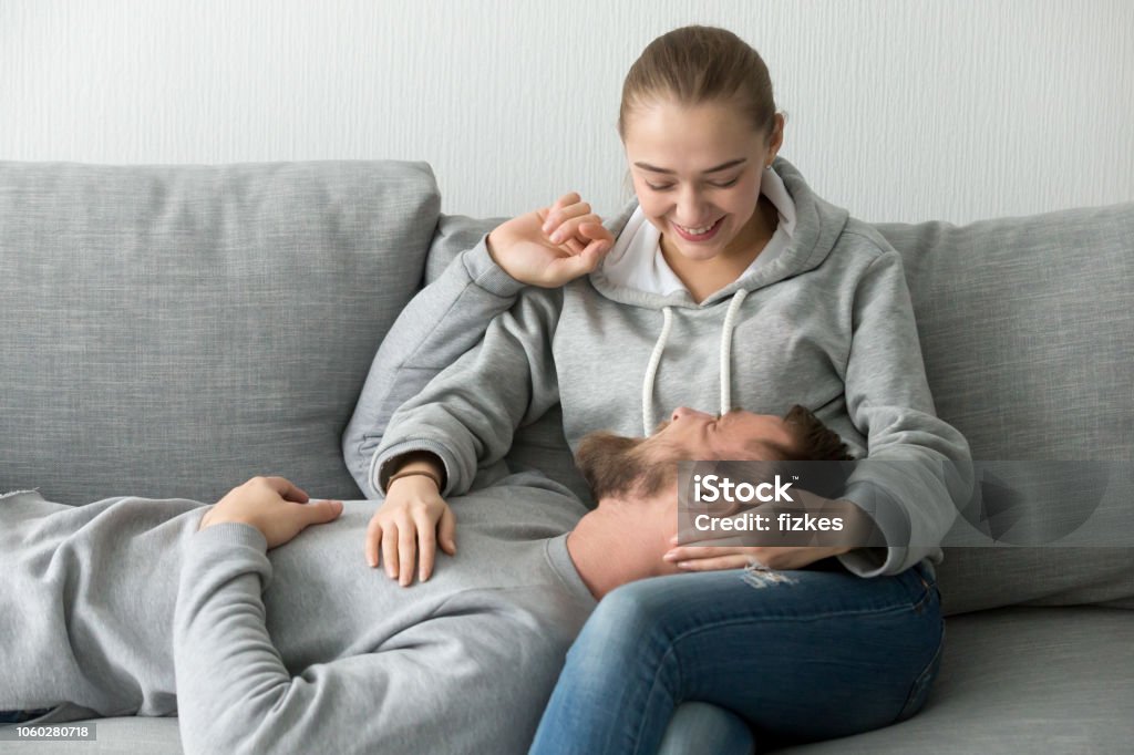 Smiling girlfriend caress lover relaxing on her knees on couch Smiling boyfriend relax on girlfriend knees on grey cozy couch, loving woman comfort and caress beloved man, laughing couple having fun resting on sofa spending weekend together at home Embracing Stock Photo