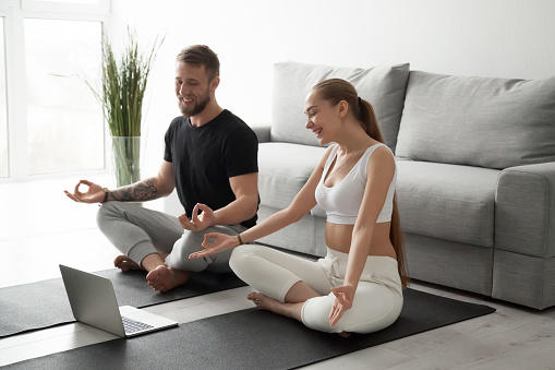 Smiling millennial couple practice yoga on mats at home watching video tutorial on laptop, excited man and woman sit in lotus position meditating repeating online instructions by coach on computer