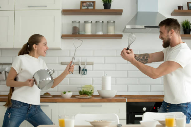 Playful young couple playing with kitchenware in kitchen Happy young couple having fun in kitchen playing with kitchenware while preparing breakfast, playful millennial man and woman cooking food at home together involved in imaginary fight with utensils dueling stock pictures, royalty-free photos & images