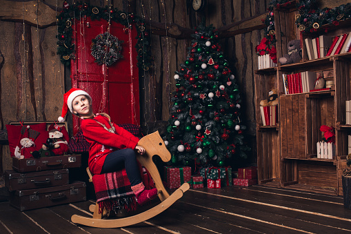 Santa girl riding rocking horse on background of Christmas decorations.