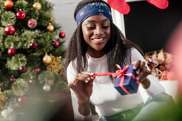 selective focus of smiling attractive african american woman opening christmas present at home selective focus of smiling attractive african american woman opening christmas present at home gift lounge stock pictures, royalty-free photos & images
