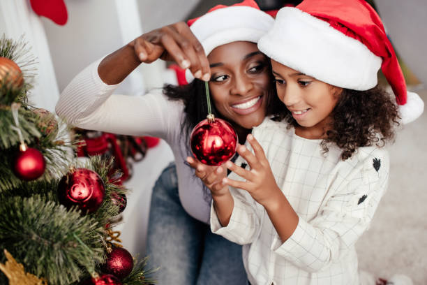 sonriente a mujer afroamericana con sombrero de santa claus ayudando a la hija para decorar árbol de navidad en casa - decor christmas celebration event christmas ornament fotografías e imágenes de stock