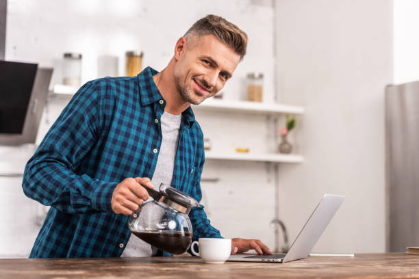 istock handsome smiling man pouring coffee and using laptop at home 1060365824