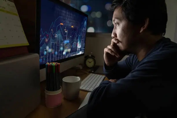 Photo of Portrait of Asia Businessman sitting and working hard on the table with Trading graph on the cityscape over front of computer screen in work place at late with serious action