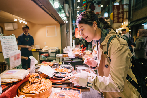 lovely female tourist standing in front of the vendor in the market and pointing to the Japanese street food. the shop owner is smiling at her in the background. Joyful backpacker travel in Japan.