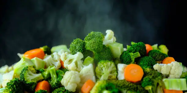 Fresh tender vibrant green steaming vegetables against a black background. Vegetables include broccoli, cauliflower, cabbage and carrots. Shallow depth of field.