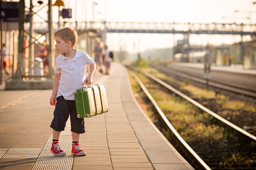 Adorable little kid boy dressed in shorts and polo t-shirt on a railway station, waiting for the train with retro old green suitcase. Ready for vacation. Young traveller on the platform.
