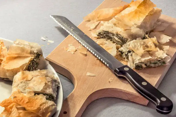 Freshly baked traditional delicious spinach cheese roll pie, sliced with serrated bread knife on wooden cutting board, and offered on porcelain platter.