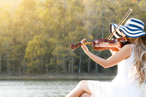 A woman / violinist wearing white dress and hat playing violin on riverside among sunlight feeling freedom