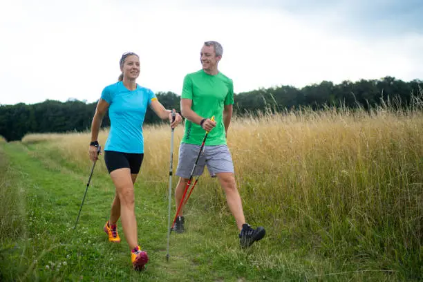 concentrated woman learning correct nordic walking technique from her personal trainer explaining during outdoor training on natural path between high grass on thundery summer afternoon