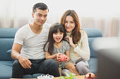 Family time young Parents and Daughter smile which sit on sofa, daughter take an apple on hand which sit between father and mother and looking TV at home