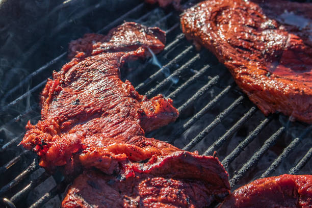Delicious marinated thin steaks grilling on outdoor cooker with fat bubbling and steam - close-up and selective focus