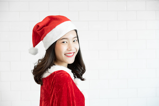 Attractive Asian Girl in Red Santa Claus dress is smiling and looking to camera on white brick wall. Copy space.