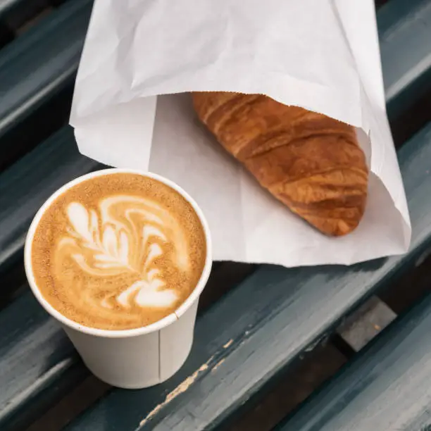 Paper cup of capupuccino coffee take-away, and croissant, on wooden bench, outdoor.