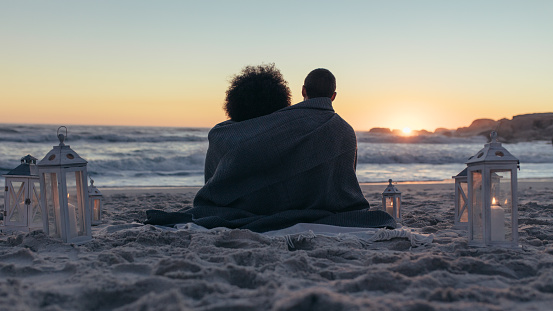 Rear view of man and woman wrapped with a blanket sitting on beach and looking at sunset. Couple admiring the sunset from sea shore.