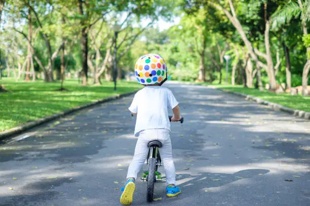 Backside of cute Asian 2 years toddler boy child wearing safety helmet learning to ride first balance bike in sunny summer day, kid cycling at park, Explore & Appreciate Nature with toddlers concept