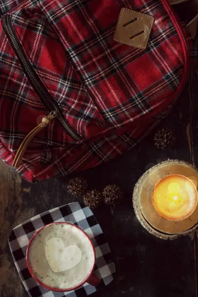 A red plaid backpack, a cup of coffee with a heart in the foam and a burning log candle, with tree seedpods scattered around. giving a study, college, holiday break feel.