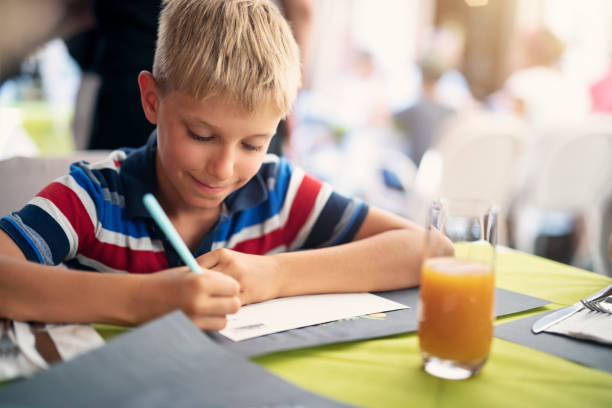 Little boy aged 9 writing postcards to friends and family. He is sitting in a street cafe in Malcesine, Italy.\nNikon D850