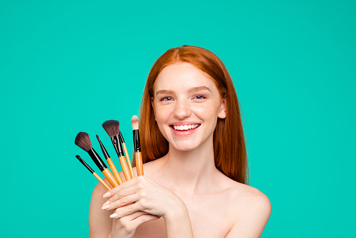 Advertising concept. Portrait of nice cheerful nude red-haired girl with shiny pure clean fresh smooth flawless skin, demonstrating brushes for cool makeup, isolated over green turquoise background