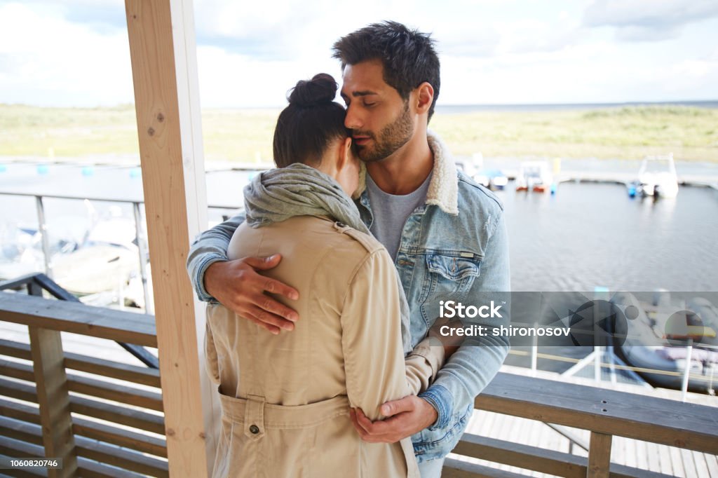 Calm loving man hugging girlfriend at seaport Serious calm loving handsome young bearded man with black hair wearing denim jacket keeping eyes closed and hugging girlfriend at seaport while consoling her Adult Stock Photo