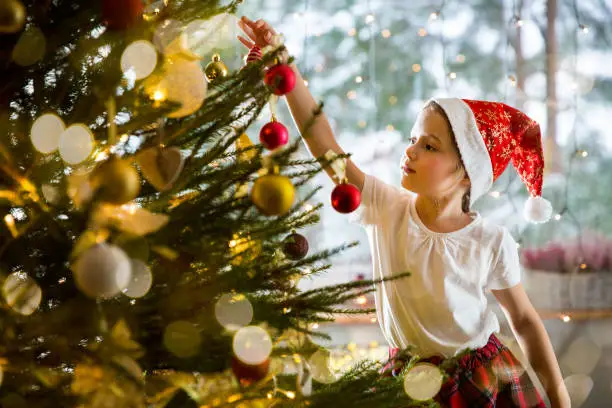 Photo of Cute little girl in red Santa hat and plaid skirt decorating Christmas tree at home.