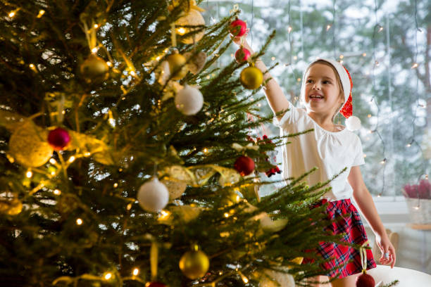 Cute little girl in red Santa hat and plaid skirt decorating Christmas tree at home. stock photo