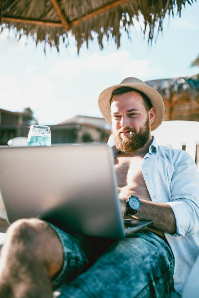 istock Bearded Hipster With Straw Hat Typing On Laptop On A Beach 1060873878