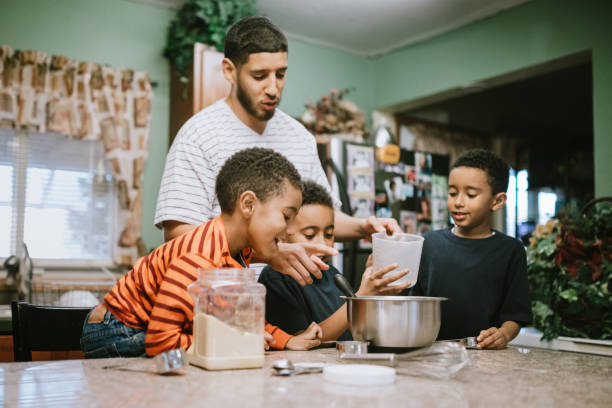 Father Prepares Meal in Kitchen With His Sons A dad and his three boys work together to make dinner at home, having fun spending family time together.  They mix and pour ingredients in to a bowl. washington state photos stock pictures, royalty-free photos & images