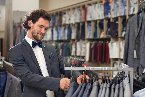 Elegant young guy searching for perfect upper part of costume. Gorgeous handsome man standing in store and trying on checkered grey and white jacket. Male jackets and shirts on background.